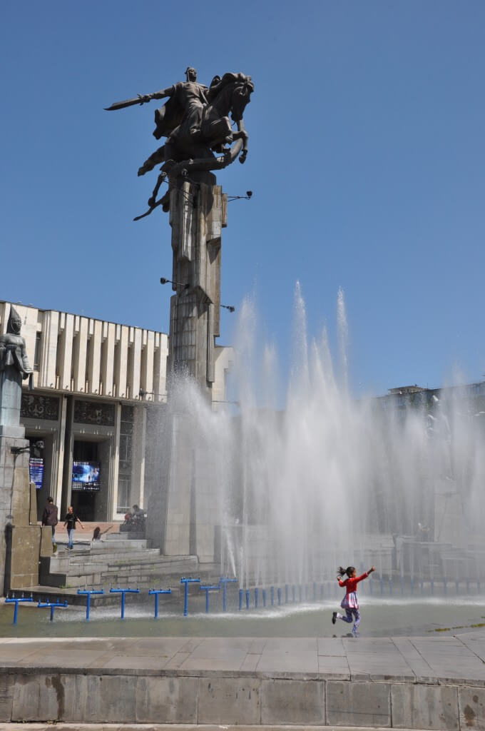 今回の旅の写真で最も気に入った一枚である、青い空とキルギスの英雄マナス像、その下で力強く噴き出す噴水と飛び跳ねる赤い服の子供